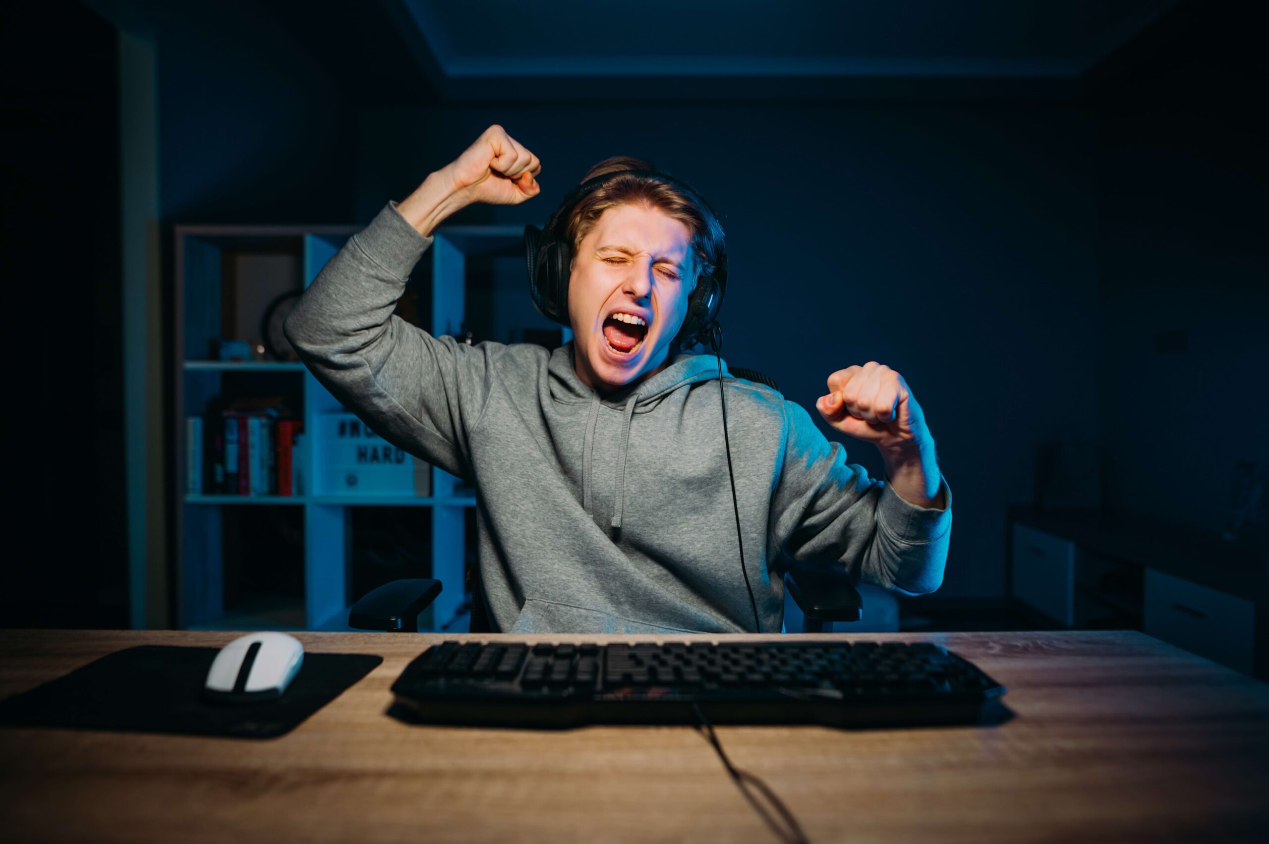 Homem jovem comemorando com sorriso e mãos levantadas no seu setup gamer