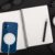 Carregador magsafe sem fio fixado em iPhone azul em cima de caderno em branco, ao lado de caneta preta, xícara de café e fones de ouvido brancos.