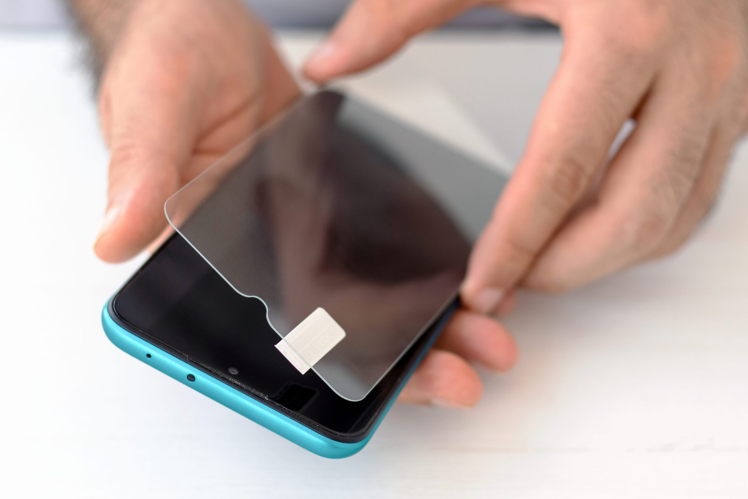 Mãos aplicando película de proteção no celular azul com a tela desligada sob uma mesa branca