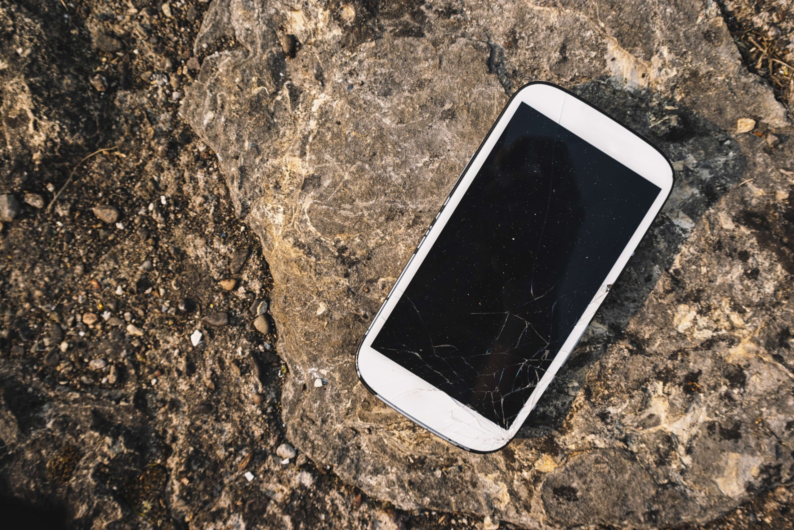 Smartphone branco com a tela quebrada no chão, em cima de uma pedra