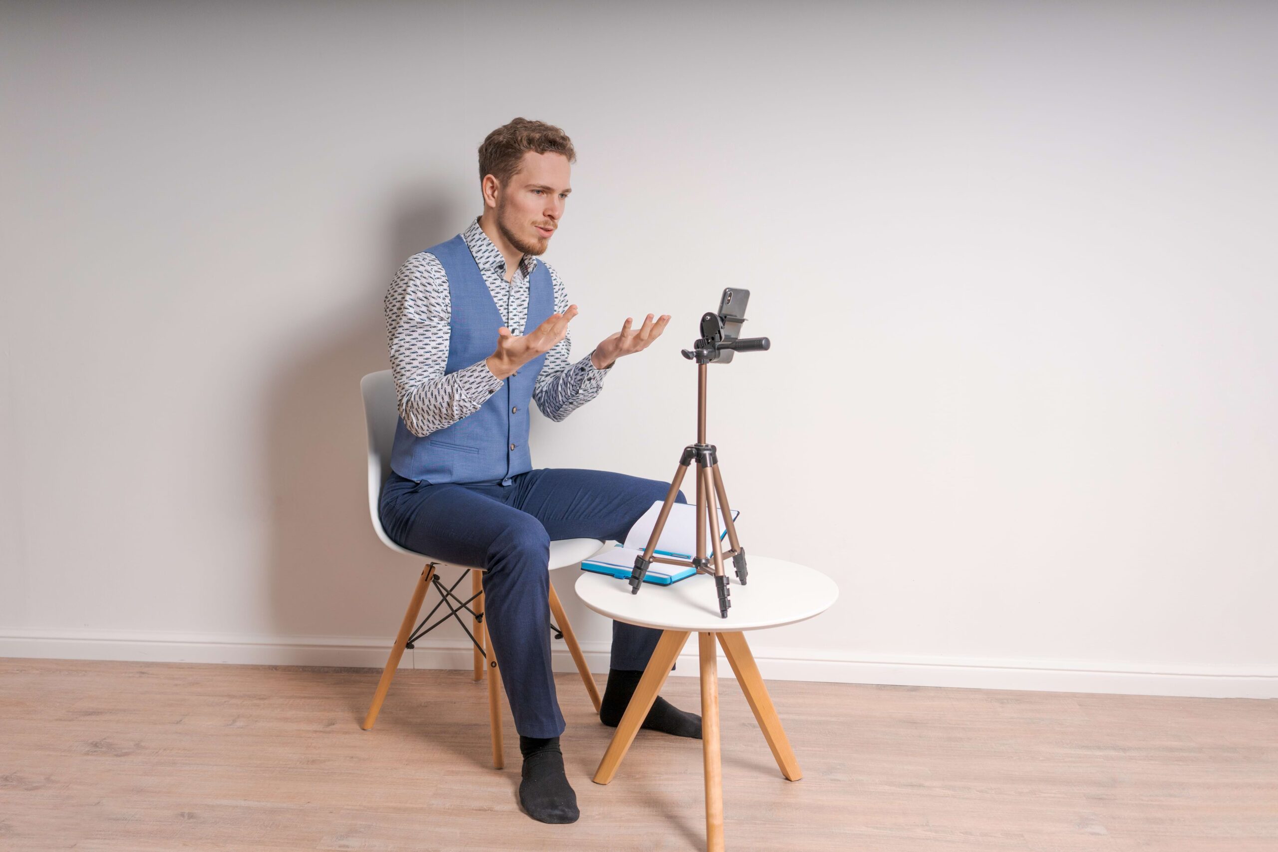 Homem sentado em cadeira branca, olhando para o celular preto em um tripé preto fazendo gestos de confusão